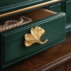 May include: A dark green wooden jewelry box with a partially open drawer. The drawer has a gold-coloured ginkgo leaf handle and contains a strand of pearls. Gold trim accents the top.
