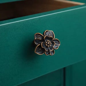 Könnte beinhalten: Ein bronzefarbener, blütenförmiger Schrankknauf ist an einer türkisgrünen Schranktür angebracht. Der Knauf hat detaillierte Blütenblätter und eine zentrale Anordnung kleiner Kreise. Die Schublade ist geöffnet und gibt einen Holzeinbau frei.