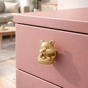 Peut inclure: Gros plan sur une commode rose avec un bouton de tiroir doré en forme de chat. Le bouton de tiroir est une sculpture détaillée d'un chat assis. La commode a une finition mate et un design simple.
