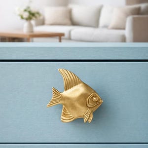 Peut inclure: Un bouton de tiroir doré en forme de poisson, monté sur un meuble bleu clair. Le poisson présente des nageoires détaillées et une surface texturée, ajoutant une touche décorative au meuble. Le meuble est peint dans une finition mate unie.