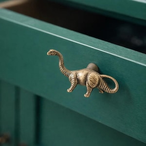 Peut inclure: Un bouton de meuble en forme de dinosaure en laiton est fixé sur une porte d'armoire vert teal. Le dinosaure a un long cou et une queue, avec une texture détaillée. La porte de l'armoire est ouverte.