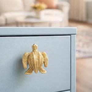 May include: A gold-coloured turtle-shaped cabinet knob is mounted on a light blue drawer. The knob is a detailed sculpture of a turtle with textured shell and flippers. The drawer is part of a larger piece of furniture.