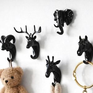 May include: A selection of black animal head wall hooks, featuring a ram, deer, elephant, horse, and giraffe. A teddy bear and a gold-coloured ring hang from the hooks. The hooks are mounted on a white wall.