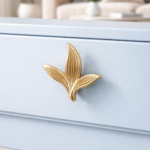 May include: A gold-colored leaf-shaped drawer pull is mounted on a light blue cabinet. The drawer pull has a detailed, textured design. The cabinet is part of a larger piece of furniture, with a blurred background.