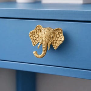 Può includere: Un pomello per cassetto a forma di elefante color oro è montato su un cassetto blu. La testa dell'elefante presenta trame dettagliate, inclusi motivi circolari sulle orecchie. Il cassetto fa parte di una scrivania o di un armadio blu.