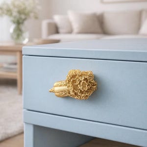 以下が含まれることがあります： 金色の装飾的な取っ手が付いた、水色の引き出し。取っ手は花の模様です。引き出しはキャビネットまたはドレッサーの一部です。背景はぼやけており、リビングルームの様子が写っています。