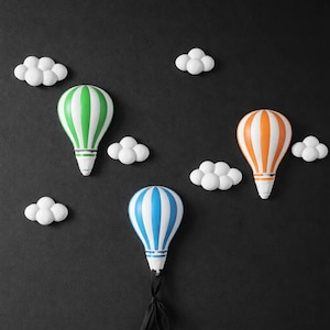 Puede incluir: Tres decoraciones de globos aerostáticos coloridos con nubes blancas sobre un fondo negro. Los globos tienen rayas verdes, naranjas y azules. Cada globo tiene una base blanca y una cinta negra.