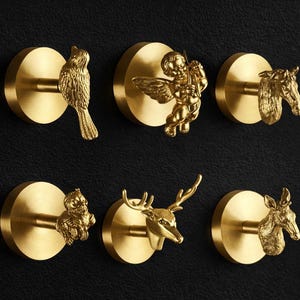 May include: A collection of six brass-colored decorative wall hooks. Each hook features a different sculpted design: a bird, an angel, a horse head, an owl, a deer head, and a donkey head. The hooks are mounted on circular backplates.