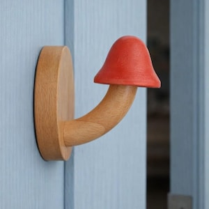 Puede incluir: Un tirador de puerta de madera con forma de seta, con un sombrero rojo y un tallo curvado. El tirador está montado en una puerta azul claro. La seta es de madera y tiene un acabado natural.