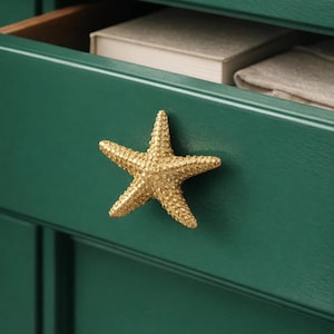 Op de afbeelding: Een gouden, stervormige knop op een donkergroene kastdeur. De lade staat gedeeltelijk open en toont een boek en gevouwen stoffen. De zeester heeft een textuur, wat een decoratieve nautische touch aan het meubel geeft.