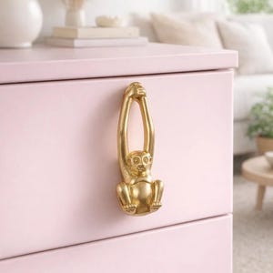 Pode incluir: Um puxador dourado em forma de macaco está preso a uma cômoda rosa. O macaco está pendurado com os braços estendidos. A cômoda tem um design limpo e moderno, com um acabamento rosa claro. O fundo inclui uma lâmpada, livros e um sofá.