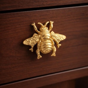 以下が含まれることがあります： 濃い茶色の木製引き出しに取り付けられた、金色のハチ型の引き出しつまみ。ハチは、詳細な羽、触角、体のセグメントを持っています。引き出しの木目が見えます。