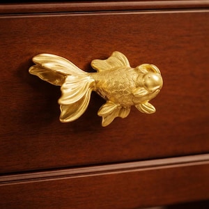 Könnte beinhalten: Ein goldfarbener, fischförmiger Schubladengriff, montiert auf einer dunkelbraunen Holzschublade. Der Griff ist eine detaillierte Skulptur eines Goldfisches, mit sichtbaren Flossen und Schuppen. Die Holzmaserung ist sichtbar.