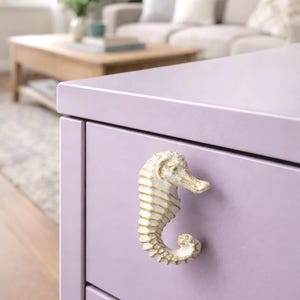 Pode incluir: Close-up de uma gaveta de cômoda lavanda com uma maçaneta em forma de cavalo-marinho. A maçaneta é branco-sujo com detalhes dourados, o cavalo-marinho está em posição vertical. O fundo é uma sala de estar desfocada.
