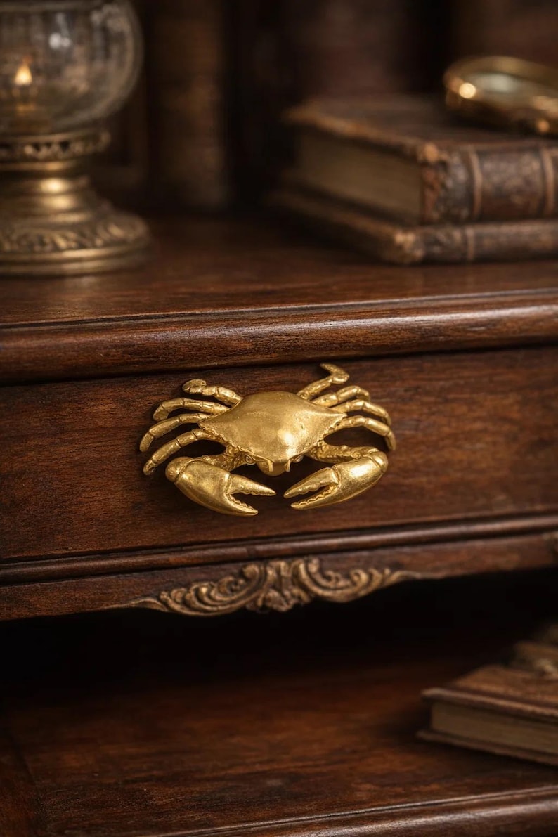 May include: A gold-colored crab-shaped drawer pull mounted on a dark brown wooden drawer. The crab has detailed claws and a textured surface. The drawer is part of a larger wooden cabinet, with other antique items in the background.
