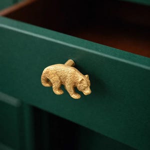 Op de afbeelding: Een goudkleurige berenvormige trekker op een donkergroene lade. De beer is gedetailleerd met vacht en een kleine kop. De lade is open en onthult een bruin interieur.