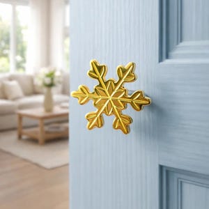 Puede incluir: Un pomo dorado en forma de copo de nieve está montado en una puerta azul claro. El pomo tiene seis puntas y un diseño detallado. La puerta forma parte de una habitación con un fondo borroso.