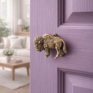 May include: A brass buffalo door knob, mounted on a lilac-painted wooden door. The knob is a detailed sculpture of a bison, adding a rustic touch to home decor. The door is part of a room with a neutral color scheme.