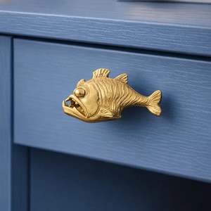 Peut inclure: Poignée de tiroir en forme de poisson de couleur laiton, fixée sur un tiroir bleu. Le poisson présente des écailles, des nageoires et une bouche ouverte avec des dents visibles. Le tiroir fait partie d'un meuble bleu.