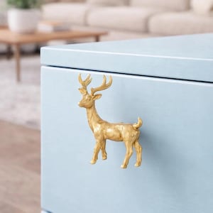 May include: A gold-colored deer-shaped cabinet knob is mounted on a light blue cabinet. The deer has detailed antlers and a textured surface. The cabinet is part of a larger piece of furniture.