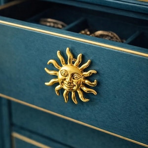 May include: Close-up of a teal jewelry box with a gold sun-shaped drawer pull. The drawer is open, revealing compartments lined with dark blue fabric. Gold trim accents the box.