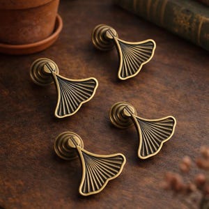 May include: Four antique brass cabinet pulls with a fan-shaped design. The pulls are arranged on a dark wooden surface, with a small potted plant and books in the background. Each pull has a diameter of approximately 1.5 inches.