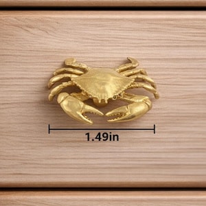 May include: A gold-colored crab-shaped cabinet pull, measuring 1.49 inches wide, is mounted on a light-colored wooden surface. The crab has detailed claws and legs, adding a nautical or coastal theme to furniture.