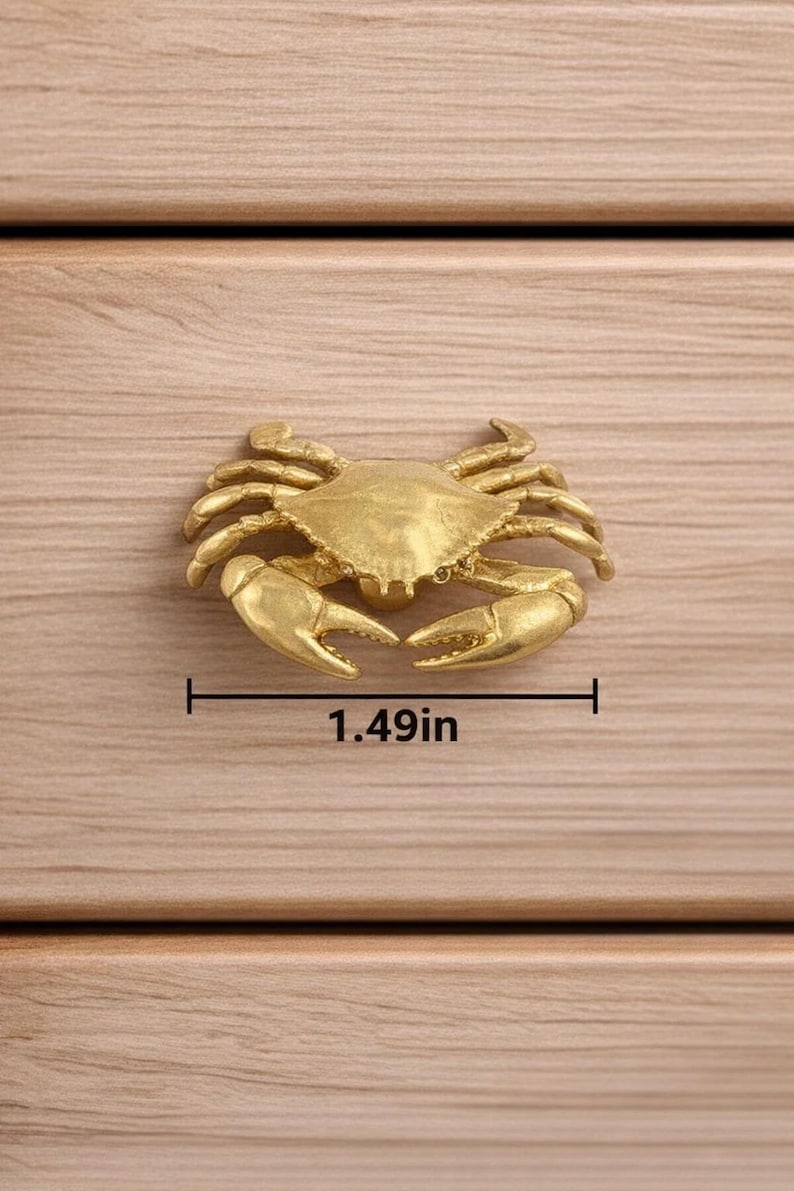 May include: A gold-colored crab-shaped cabinet pull, measuring 1.49 inches wide, is mounted on a light-colored wooden surface. The crab has detailed claws and legs, adding a nautical or coastal theme to furniture.