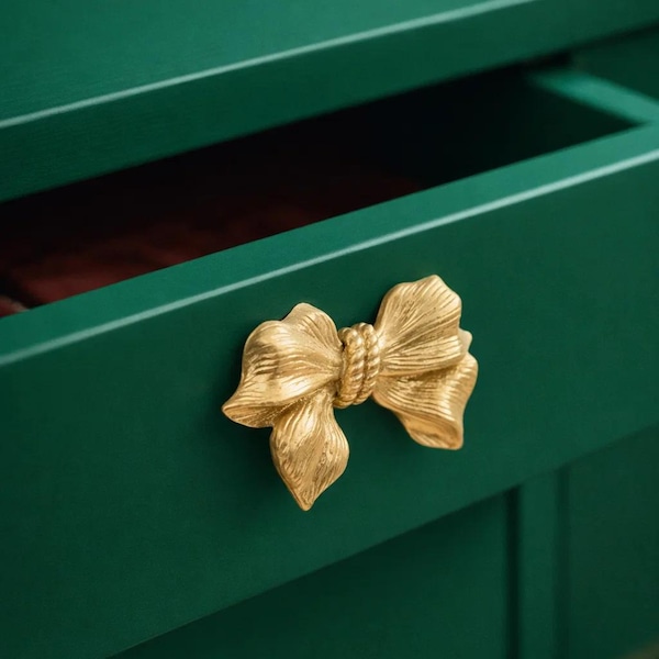 May include: Close-up of a green drawer with a gold bow-shaped handle. The drawer is open, revealing a glimpse of the interior. The bow handle adds a decorative touch to the furniture.