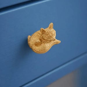 Könnte beinhalten: Ein goldfarbener, fuchsförmiger Schubladengriff ist an einem blauen Schrank befestigt. Der Fuchs ist zusammengerollt, wobei sein Schwanz den Griff bildet. Der Schrank hat eine glatte, lackierte Oberfläche.