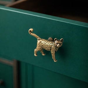 Op de afbeelding: Een messing katvormige ladeknop is bevestigd op een teal groene lade. De kat wordt afgebeeld in een loopstand met zijn staart gekruld. De lade maakt deel uit van een groter meubelstuk, met een open lade en een donker interieur zichtbaar.