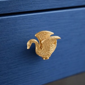 May include: A gold swan-shaped drawer pull, mounted on a blue wooden surface. The swan has outstretched wings and a curved neck. This decorative hardware element is designed for furniture such as drawers or cabinets.