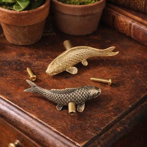 May include: Two decorative fish-shaped cabinet pulls. One is gold-toned, the other is silver-toned. Both have detailed scale patterns and are mounted on brass posts. The pulls are displayed on a dark wooden surface, suggesting a vintage or antique aesthetic.