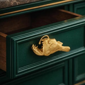 Puede incluir: Cajón de armario verde oscuro con un tirador dorado en forma de pez rape. El cajón está abierto, revelando un interior de madera. El armario tiene una tapa de mármol y ribetes dorados.