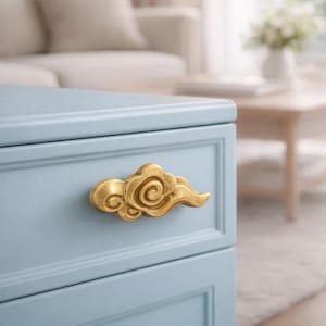 Może przedstawiać: Zbliżenie jasnoniebieskiej szafki ze złotą, chmurową gałką szuflady. Gałka szuflady ma szczegółowy, wirujący wzór. Szafka znajduje się w pomieszczeniu o neutralnej palecie kolorów.