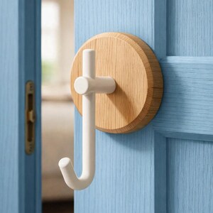 Puede incluir: Un gancho blanco en forma de J con una barra transversal, montado sobre una base redonda de madera clara. El gancho está fijado a una puerta azul claro, creando un diseño minimalista. El gancho está diseñado para colgar objetos.