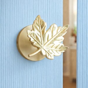 May include: A gold-coloured maple leaf door knob. The knob is mounted on a round base and is set against a light blue door. The leaf design has detailed veining, adding to its realistic appearance. The door knob is a decorative hardware item.