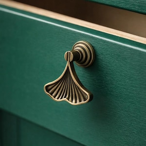Peut inclure: Gros plan sur une poignée de meuble en bronze en forme de feuille de ginkgo. La poignée est fixée sur une porte d'armoire vert foncé. Le tiroir est ouvert, révélant un intérieur clair.