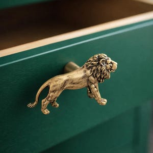 May include: A gold-colored lion-shaped drawer pull on a dark green drawer. The lion is in a walking pose, with detailed mane and body. The drawer is open, revealing a wooden interior.