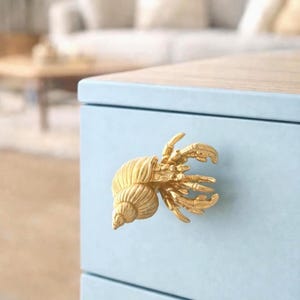 Pode incluir: Puxador de armário dourado em forma de caranguejo eremita. O puxador está fixado a uma gaveta de armário azul claro. O fundo está desfocado, mostrando uma sala com um sofá e uma mesa de madeira.