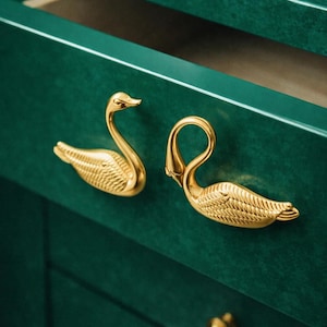 May include: Close-up of a dark green cabinet drawer with two gold swan-shaped drawer pulls. The swan pulls are detailed with textured feathers and curved necks. A small gold knob is visible below.