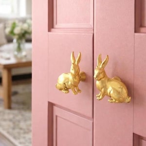 Op de afbeelding: Twee goudkleurige konijnvormige kastknoppen gemonteerd op een roze kastdeur. De knoppen hebben rechtopstaande oren en een zittende pose. De kastdeur is geschilderd in een zachte roze kleur.