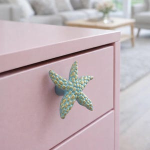 Pode incluir: Uma cômoda rosa claro com um puxador em forma de estrela do mar. O puxador é azul com detalhes dourados. A cômoda está em uma sala com um sofá e um tapete de cor clara.
