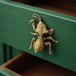 Può includere: Pomello per cassetto a forma di insetto in ottone dettagliato montato su un frontale di cassetto verniciato di verde. Il cassetto è aperto, rivelando un interno in legno. Ferramenta decorativa.