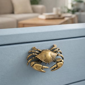 May include: A brass crab-shaped cabinet pull is mounted on a light blue drawer. The crab has detailed claws and a textured shell. The drawer is part of a larger piece of furniture, with a blurred background.