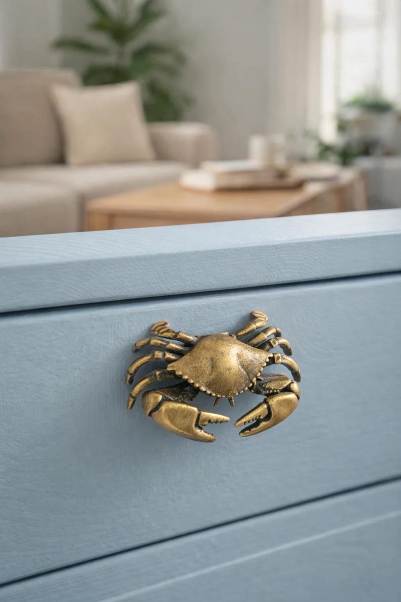 May include: A brass crab-shaped cabinet pull is mounted on a light blue drawer. The crab has detailed claws and a textured shell. The drawer is part of a larger piece of furniture, with a blurred background.