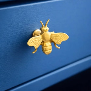 Puede incluir: Un tirador de cajón dorado con forma de abeja sobre una superficie azul. La abeja tiene alas, antenas y segmentos corporales detallados. Este herraje decorativo añade un toque de elegancia inspirada en la naturaleza a los muebles.