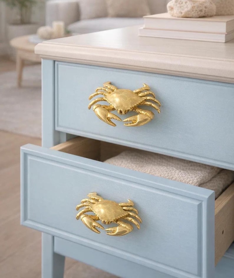 May include: A light blue bedside table with two drawers, each adorned with a gold-colored crab-shaped handle. The top of the table is a light beige color. One drawer is open, revealing a folded beige blanket.