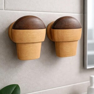 Puede incluir: Dos perchas de pared de madera con un diseño bicolor. La parte superior es de color marrón oscuro y redondeada, y la base es de madera clara en forma de cono. Están montadas en una pared de azulejos gris claro, ideal para un baño.