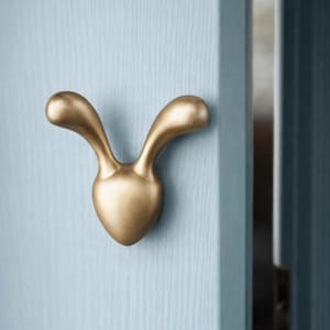 May include: A gold-colored metal hook shaped like a rabbit's head is mounted on a light blue wooden door. The hook has a smooth, rounded design with two upward-curving ears and a pointed base. The door has a vertical wood grain texture.